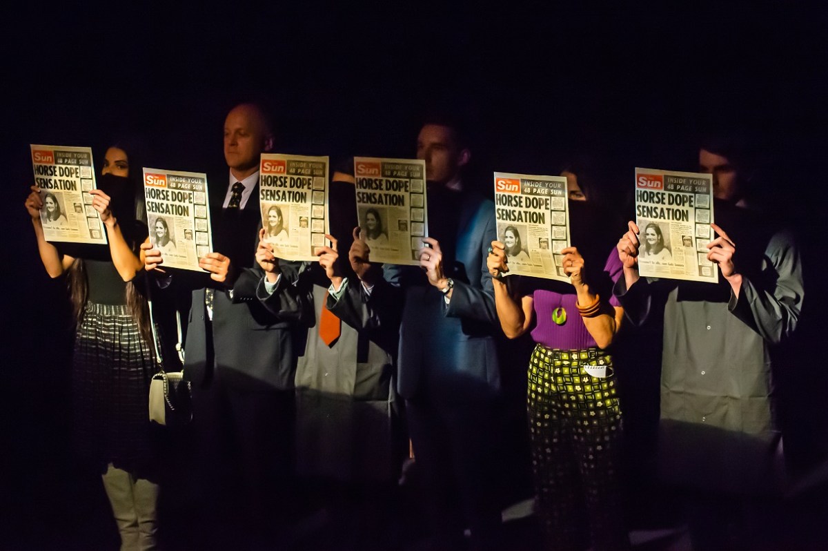 Ink by New Theatre. Six people are lined up on a dark stage holding identical copies of The Sun newspaper in front of themselves, obscuring their faces.