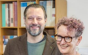 A bearded man with a green top and brown jacket is standing next to a woman with glasses and short curly hair. There is a book case behind them. They are both smiling.