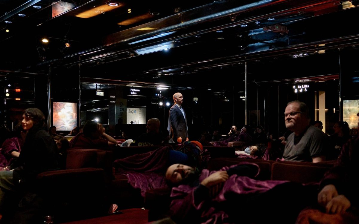 A figure stands in a dark underground space with sofas laid out and people sleeping.