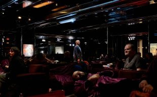 A figure stands in a dark underground space with sofas laid out and people sleeping.