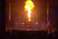 A woman is kneeling in the middle of a stage, with her head back. Plumes of fire are coming out of her mouth
