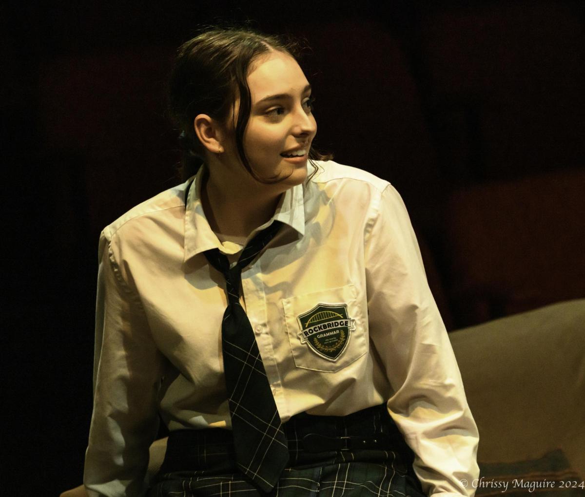 A teenage girl with her brunette hair in a ponytail is wearing school uniform and looking over to the side.