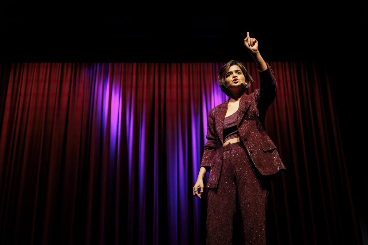 Suhani Shah. A woman with a short bob haircut wearing a sparkling maroon pantsuit is standing on stage in front of heavy curtains. She has her left arm raised and fingers pointed to the sky.