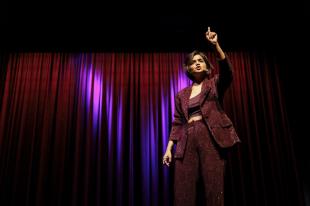 Suhani Shah. A woman with a short bob haircut wearing a sparkling maroon pantsuit is standing on stage in front of heavy curtains. She has her left arm raised and fingers pointed to the sky.