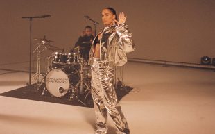 A woman in a silver top and trousers is standing in front of a drummer and his drum kit.