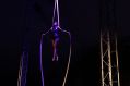A female acrobat hanging from silk ropes at height in front of stage curtains.