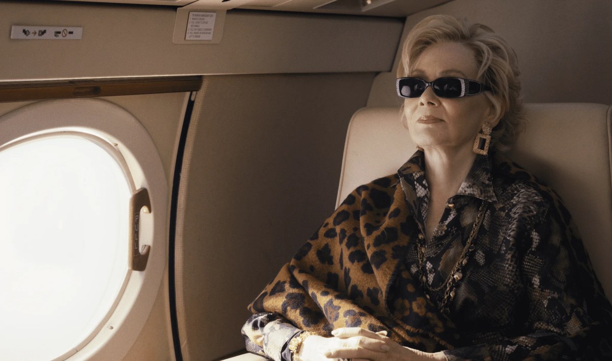An older woman wearing dark glasses, gold jewellery, expensive clothes and an enigmatic expression sits back in her seat in a private jet.