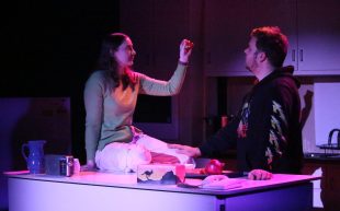 Julia Slater-Allan and Sam Corr in ‘Cutting Onions’. Photo: Supplied. A woman sitting on the countertop based in purple light in a domestic sitting, gesturing to a man wearing a black hoodie.
