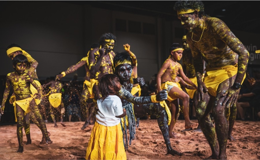 Peppimenarti Dancers, Darwin Aboriginal Art Fair 2023. Photo: Tamati Smith. Aboriginal dancers in yellow body paint are interacting with a young child in their midst. They are stepping on red sand inside a convention centre. 