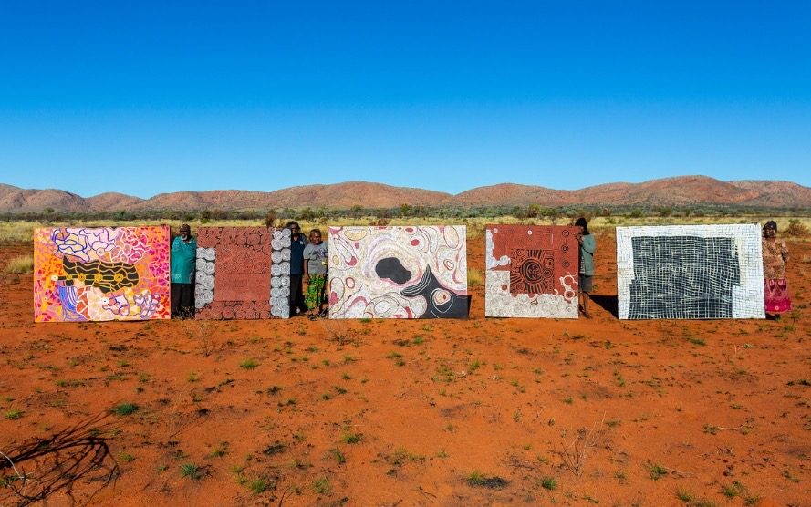 Ngaanyatjarra Land, artists from L-R: Nyungawarra Ward, Dorcas Tinamayi Bennett, Cynthia Burke, Delilah Shepherd and Nancy Nyanyarna Jackson. Photo: Jason Thomas. Image: Courtesy of Warakurna Artists. Five large-scale paintings held up by artists in the Australian desert landscape with a blue skyline in the background.