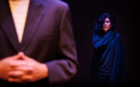 Two figures stand on a dark stage, one is wearing a suit jacket and very close to the camera with only the upper body visible. Another stands further back, with curly black hair, black clothing and a solemn expression.