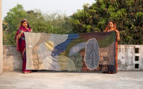 Two people hold up a large textile work in an outdoor setting.