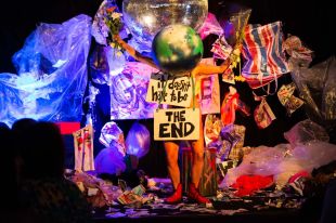 Festival of Dangerous Ideas return to Carriageworks. Photo: Courtesy of FODI. A chaotic stage with a person wearing an earth globe on its head and the signs ‘it doesn’t have to be THE END’.