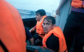 Photo of asylum seekers captured by Barat Ali Batoor, in ‘Searching for Photo courtesy of Barat Ali Batoor. The composition of the photo has highlighted two asylum seekers wearing orange life jackets on a boat. One is looking towards the camera, his hair wet, and eyes round.