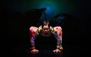 Sprung Ensemble member, Sinead Skorka Brennan in ‘O, How I Dreamt of Things Impossible’, 2020. Photo: Kate Holmes. A performer on a darkened stage with a colourful bodysuit. The performer lowered their body, supported by their arms and legs like a spider pose.