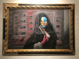 Rhodes. Image is a surreal piece of art in a golden frame, a picture of a young woman in a black cloak holding a closed fan in a red gloved hand. Her face is painted blue, yellow and white and there is a perspex or glass box around her head. She is standing in front of a block of flats.