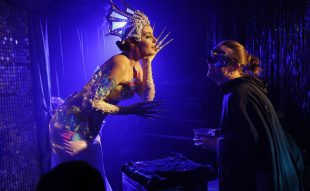 The lighting is blue. On the left a woman dressed up in metallic silver with matching headgear and long sharp nails is looking at a man in a black cloak and a mask on the right.
