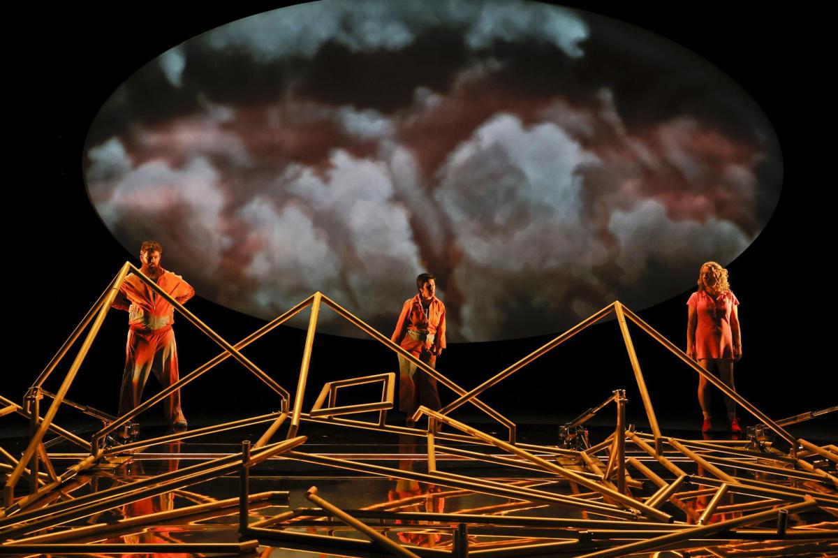 Three actors in orange hi-vis clothes with a huge oval backdrop showing clouds, stand behind a mass of complicated poles.