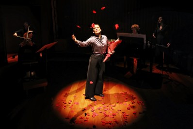A woman with her hair in a bun is standing spotlit in centre of stage. She is wearing silver blouse and black pants and carrying a bouquet of roses. Petals are strewn below her. A band can be seen playing around her, in semi darkness.