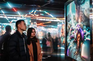 Two young people (in their twenties) are looking at a large screen image of a colourful AI-generated artwork in a darkened museum gallery space.