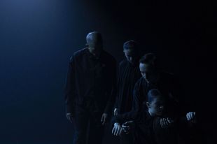 A darkened stage with four performers dressed in black in a simple formation, some are on the floor, others are standing.