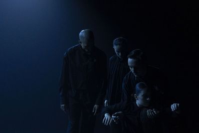 A darkened stage with four performers dressed in black in a simple formation, some are on the floor, others are standing.
