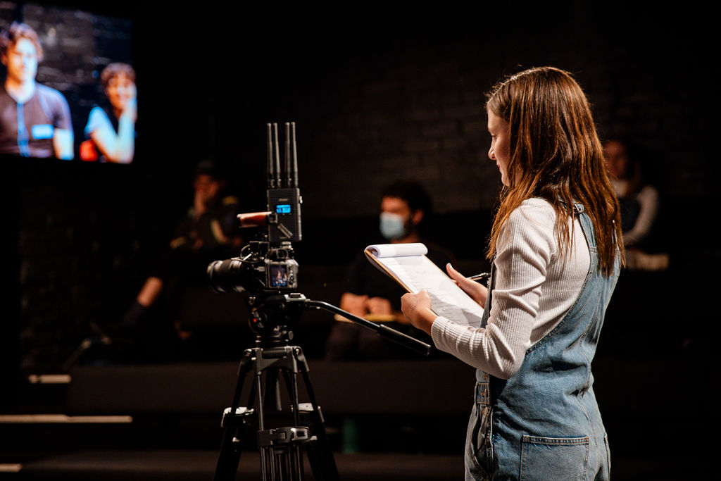 Right side profile of teenage girl who's reading a script. She's facing a camera. A projected image of a man and a woman is presented on a screen above on the left hand side.