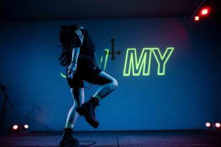 A person in shorts and boots has one knee raised in the air while dancing with their head thrown back against a blue background. The word MY can be seen in green neon behind them.