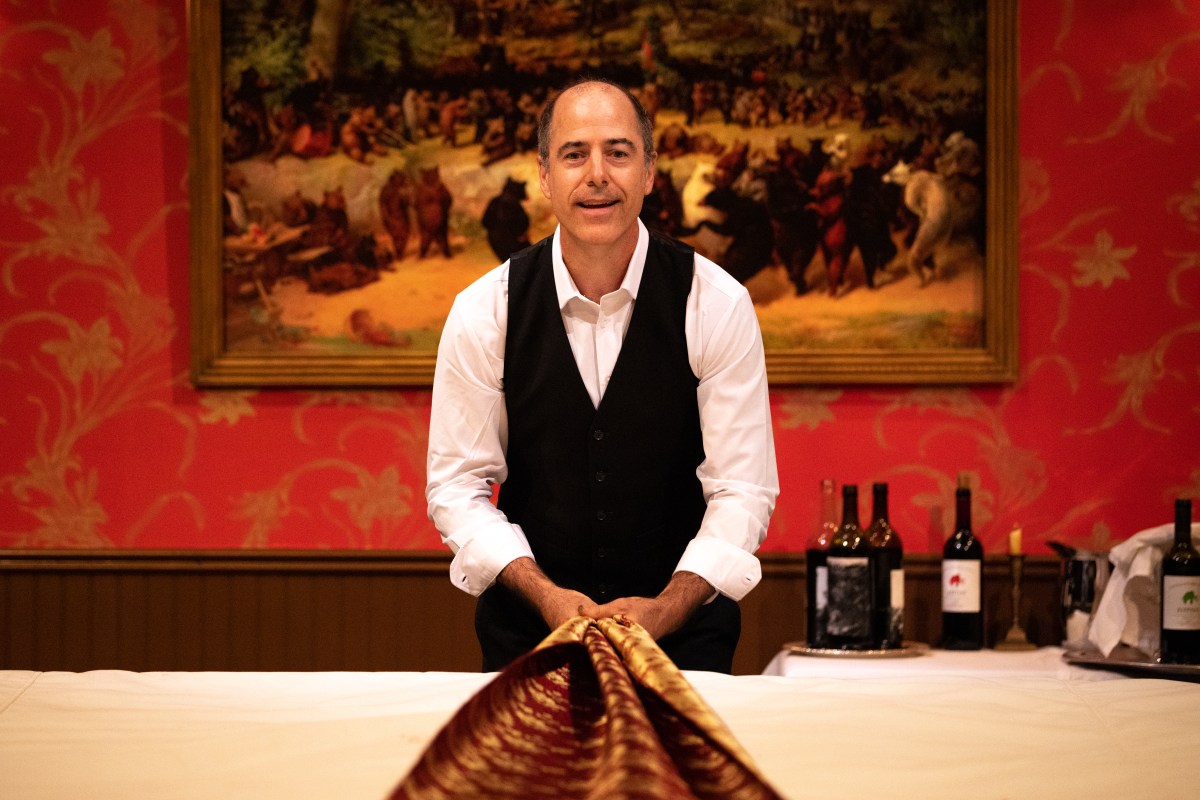 RISING: A man dressed as a waiter is smiling at the camera and pulling a table runner towards himself. Behind him is a detailed painting of anthropomorphic cats on the wall and some wine bottles.