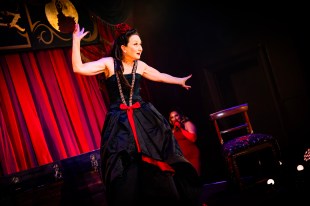 A woman is standing in front of a red curtain drape. She has dark hair and is dressed in black, with red trim. She is gesticulating, with arms stretched out, dramatically.