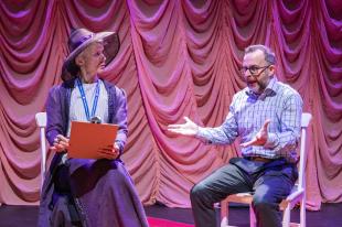 Against pink swirly drapes, a man and a woman are sitting on chairs. She on the left is dressed in an old fashioned dress and a wide brimmed hat and carrying a clipboard. He is wearing modern clothes: pants and a shirt. He has his arms wide, in the middle of talking.