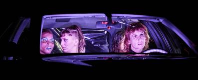 Close up of four women sitting in a car at night; they are lit from inside the car.