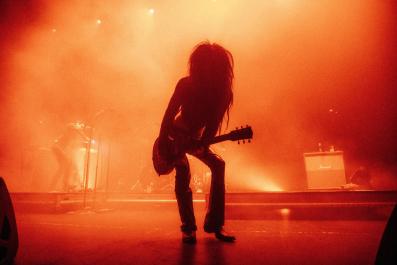 A silhouette of a figure in black on a stage holding a guitar. Behind them there is an orange haze.