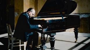 A Caucasian man is wearing a black suit and playing on a black grand piano.