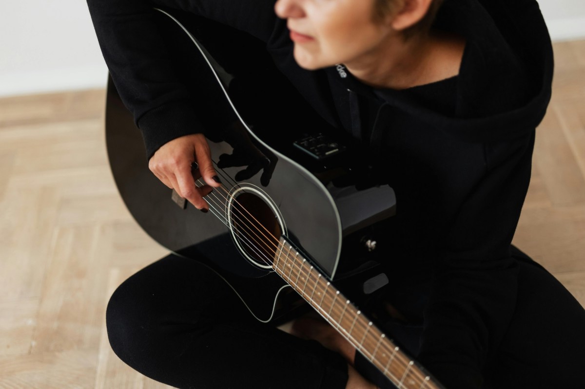 Woman dressed in black seated on floor playing black guitar.