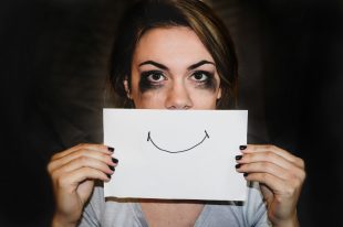 Creative burnout is common and profoundly impact your health. In this picture, a woman with stained mascara holds up a smiley face to cover her sadness.