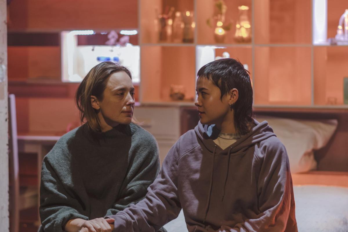 Two young women are seated and looking at each other, hand in hand. The one on the left is fair, the one of the right is brunette. Behind them is shelving with a collection of glass containers.