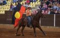 Mount Isa Mines Indigenous Rodeo Championships brings together riders and artists. A First nations man riding on a horse inside rodeo grounds while holding an Aboriginal flag.
