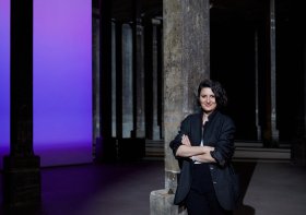 Woman with dark hair and dressed in black in dark gallery with purple illumination in background. Angelica Mesiti. Rite of When.