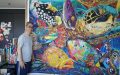 Textile artist Danny Amazonas standing in front of his large-scale quilt work. Photo: Courtesy of the artist. A middle-aged Asian man wearing glasses with yellow lenses and a gray t-shirt stnading in front of a large quilt work depicting a seaturtle surrounded by fish. The piece is vibrant and colourful.