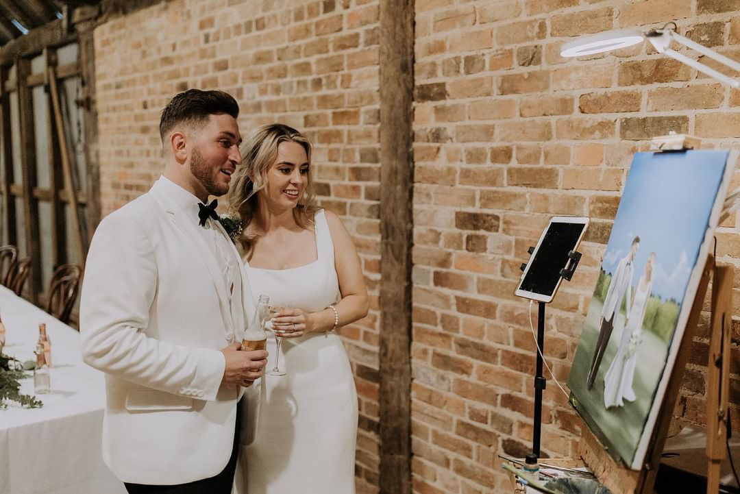 Newlyweds looking at a live wedding painting by live wedding painter Rebekka Lord-Johnson. A heterosexual couple dressed in white holding drinks while admiring a painting of them, displaying on an easel. 