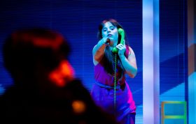 On a stage lit by a blue wash and with slat blinds as the backdrop, a young woman holding a telephone receiver to her ear points towards an audience member who can be seen from behind in silhouette. Alter State.