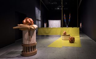 'Future Remains: The 2024 Macfarlane Commissions', installation view at ACCA. A darkened exhibition space filled by an installation towards the foreground of the photo. It features terracotta sculptures on wooden plinths made with discarded material.