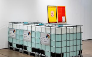 'Duty of Care' installation view at IMA. Featuring three industrial sized tubs filled with an aqua liquid and a yellow and red poster diagonally behind the tubs.