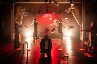 A man in shadow is crouched over in the centre of the room. Around him are four people, two standing on chairs. They are throwing bit of paper in the air. The lighting is awash in red and white spotlight. Elegies: A Song Cycle.