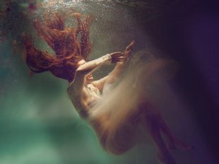 A stylised photograph of a naked woman sinking underwater, he long red hair drifting towards the surface.
