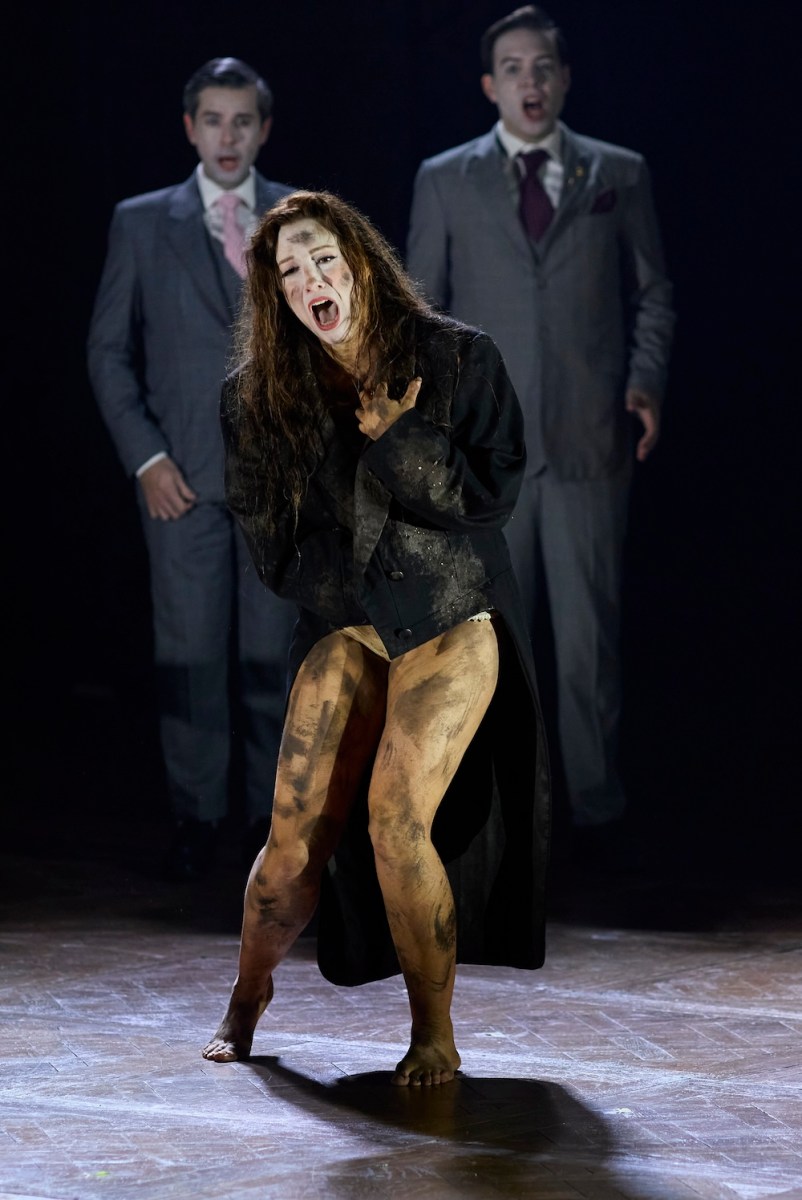 Hamlet. A mud spattered woman sings and cringes, wearing a black jacket and no trousers. Behind her are two men in grey suits.