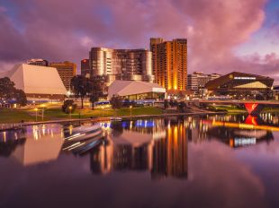 The angular buildings of Adelaide Festival Centre sit on the banks of the River Torrens, surrounded by parkland and taller buildings in the background. The photo is taken at the golden hour in late afternoon, and the buildings are blurrily reflected in the river.