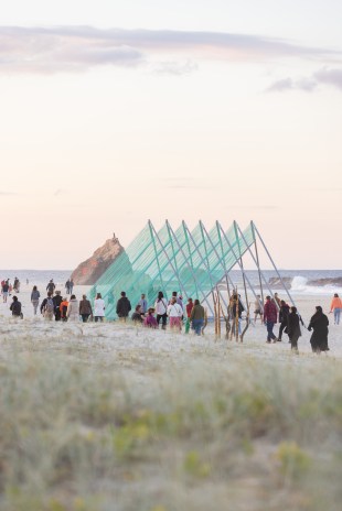 Swell Festival. On a beach a large sculpture comprising metal triangles and attached blue cords is in the centre of the frame. Behind is the sea and a rock island. People are milling about looking at the sculpture.