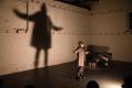 A man on a dimly lit stage has his arms outstretched. His large shadow is projected behind him on the wall. An Imaginary Life.